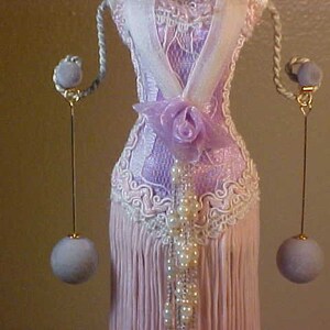 May include: A pair of long, dangling earrings with a light purple ball at the end of each earring. The earrings are hanging from a gold-colored hook and are attached to a white lace dress form.