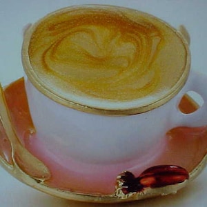 May include: A white teacup and saucer with a gold rim. The teacup is filled with a light brown liquid with a swirl pattern. The saucer is pink with a gold rim and a red and gold decorative accent.