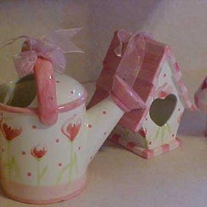 May include: A set of three pink and white ceramic home decor items. The items are a watering can, a birdhouse, and a small vase. The watering can has a pink handle and a spout. The birdhouse has a heart-shaped opening. The vase is round and has a pink rim.