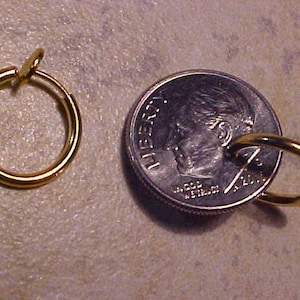 May include: A gold-toned clip-on earring with a circular design. The earring is shown next to a US dime for size comparison.