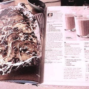 K&ouml;nnte beinhalten: Ein offenes Kochbuch zeigt ein Rezept mit drei Gl&auml;sern eines braunen Getr&auml;nks. Auf der gegen&uuml;berliegenden Seite befindet sich ein gebackenes Gericht in Folie, das mit dunklen Zutaten belegt ist. Eine kleine Sch&uuml;ssel und ein Spatel sind ebenfalls sichtbar.