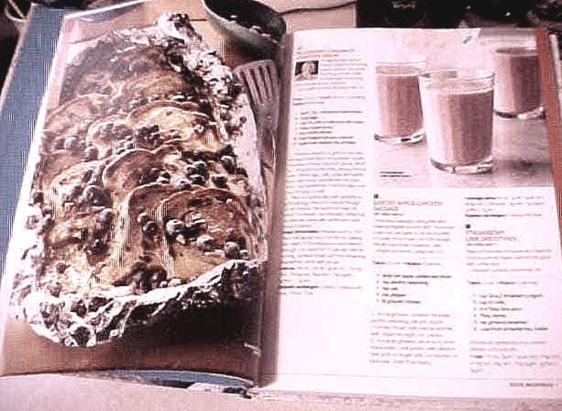 K&ouml;nnte beinhalten: Ein offenes Kochbuch zeigt ein Rezept mit drei Gl&auml;sern eines braunen Getr&auml;nks. Auf der gegen&uuml;berliegenden Seite befindet sich ein gebackenes Gericht in Folie, das mit dunklen Zutaten belegt ist. Eine kleine Sch&uuml;ssel und ein Spatel sind ebenfalls sichtbar.