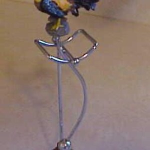 May include: A ceramic rooster figurine perched on a metal stand with a red barn base. The rooster is yellow, red, and black with a black tail.