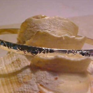 May include: A silver bangle bracelet with an engraved floral pattern. The bracelet is displayed on a light-colored, textured surface, possibly a seashell. The bangle's design is visible, reflecting light.