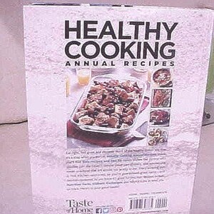 K&ouml;nnte beinhalten: Ein Kochbuch mit dem Titel "Healthy Cooking Annual Recipes" mit helllila Hintergrund. Das Cover zeigt den Titel in gro&szlig;en schwarzen Buchstaben, ein Foto eines Auflaufs und mehrere kleinere Essensbilder. Das Taste of Home-Logo befindet sich unten.