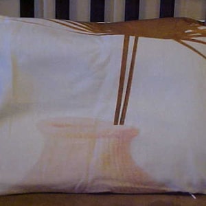 May include: A rectangular pillow with a white background and a watercolor-style design. The design features a light pink vase with brown stems and a brown, abstract design at the top. The pillow is on a brown surface.