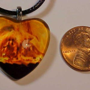 May include: Heart-shaped pendant with a fiery design in shades of orange, yellow, and black, suspended from a black cord. A U.S. penny is next to the pendant for size comparison. The pendant has a glossy finish.