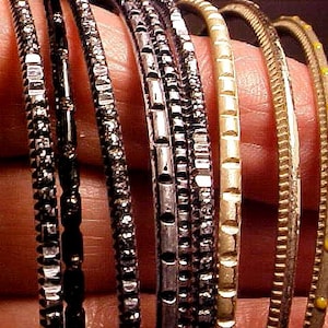 May include: A collection of seven thin bangles featuring varied textures and finishes. The set includes dark metallic bands with faceted surfaces, a smooth gold-toned band, and two lighter metallic bands, one featuring small yellow dot accents.