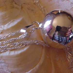 May include: A silver charm necklace with a large, round, reflective silver charm. The charm is smooth and shiny, and it reflects the light.
