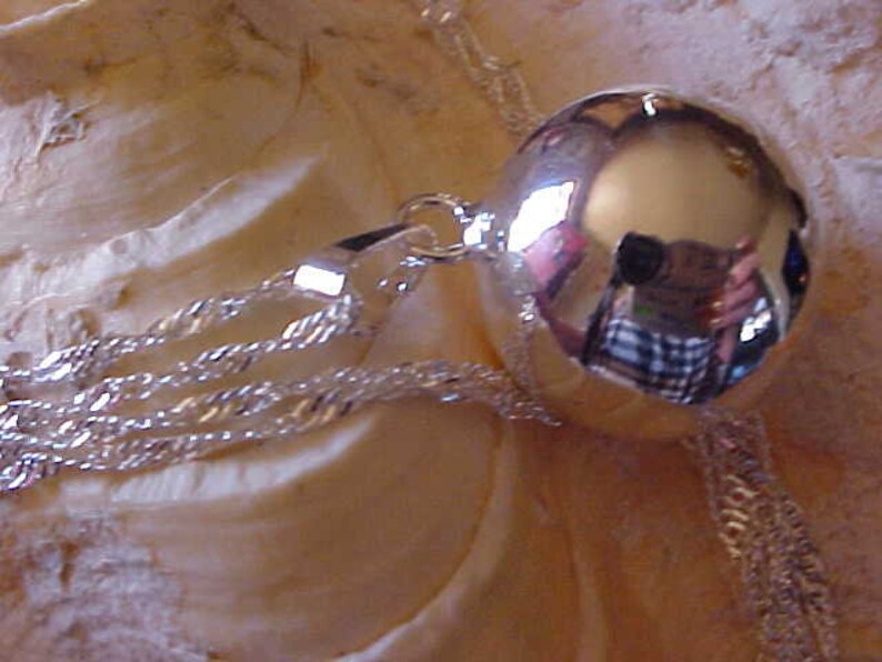 May include: A silver charm necklace with a large, round, reflective silver charm. The charm is smooth and shiny, and it reflects the light.