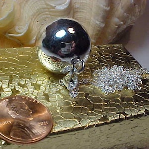 May include: A silver charm necklace with a round, silver charm. The charm is smooth and shiny. The necklace is on a gold-colored box with a textured surface.