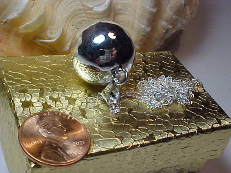 May include: A silver charm necklace with a round, silver charm. The charm is smooth and shiny. The necklace is on a gold-colored box with a textured surface.