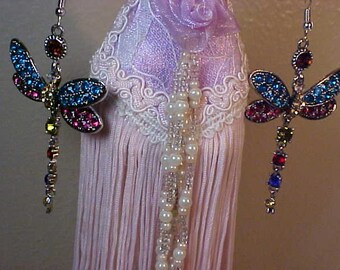 Puede incluir: Un par de pendientes de libélula plateados. Cada pendiente presenta un diseño de libélula con detalles de cristal azul, rosa y rojo en las alas. Los pendientes tienen una hebra colgante de cristales de colores y un pequeño cristal redondo en la parte superior.