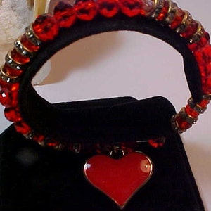May include: A red beaded bracelet with a large red heart charm. The bracelet is made of small red glass beads and gold-toned metal beads.