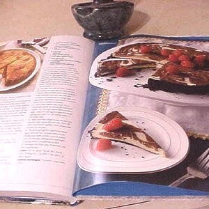 K&ouml;nnte beinhalten: Kochbuch mit Dessertbildern ge&ouml;ffnet. Eine Seite zeigt einen geschnittenen Kuchen mit Beeren, eine andere einen ganzen Kuchen mit einem entfernten St&uuml;ck. Ein kleiner, dunkler Steinm&ouml;rser befindet sich im Hintergrund.