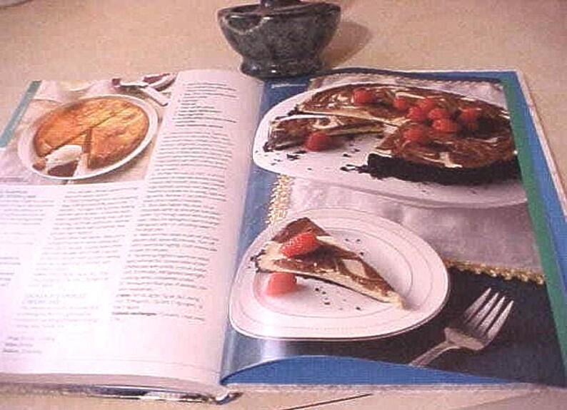K&ouml;nnte beinhalten: Kochbuch mit Dessertbildern ge&ouml;ffnet. Eine Seite zeigt einen geschnittenen Kuchen mit Beeren, eine andere einen ganzen Kuchen mit einem entfernten St&uuml;ck. Ein kleiner, dunkler Steinm&ouml;rser befindet sich im Hintergrund.