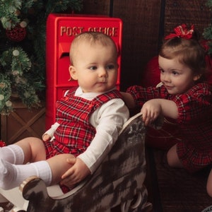 May include: Two toddlers wearing red and black plaid outfits pose for a Christmas photo. One child sits in a wooden sleigh while the other child reaches out to touch the sleigh. A red post office box is in the background.