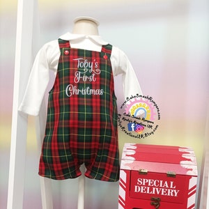 May include: A red and green tartan romper with the text "Toby's First Christmas" embroidered on the front. The romper is on a white mannequin and next to a red post box with the text "Special Delivery Santa Mail Christmas Delivery".