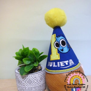 May include: Blue and yellow birthday hat with a number one and a cartoon fish on it. The hat says "Julieta" on the bottom.