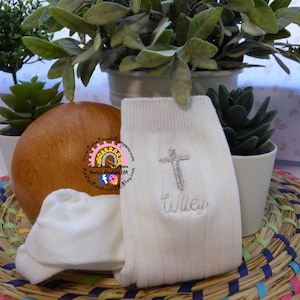 Può includere: Calzini bianchi a maglia a coste con una croce argentata e il nome "Wiley" ricamato con filo d'argento. Un calzino bianco piccolo è accanto al calzino più grande. I calzini sono esposti con un oggetto di legno e piante.