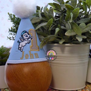 May include: A light blue birthday party hat with a white pom-pom, embroidered with a winged creature and the number 1. The name "John" is embroidered below the number. The hat is displayed on a wooden form.