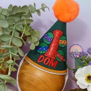 Puede incluir: Gorro de fiesta verde con un pompón naranja, con caras de las Tortugas Ninja y el número "1" en purpurina roja. El nombre "DOM" está bordado en el gorro, ideal para un primer cumpleaños.