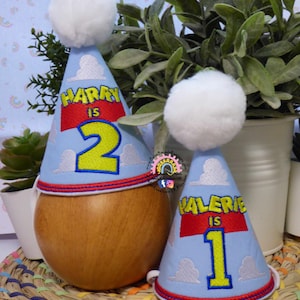 Könnte beinhalten: Zwei hellblaue Partyhüte mit weißen Wolken und einem weißen Bommel. Einer sagt "HARRY IS 2", der andere "VALERIE IS 1". Beide haben ein rotes Band mit gelben Buchstaben und einen roten Rand.