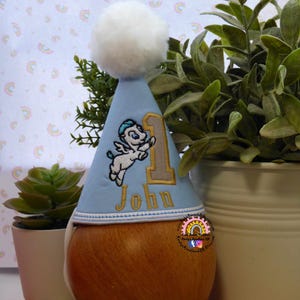 May include: A light blue birthday party hat with a white pom-pom on top. The hat features an embroidered design of a winged horse, the number 1, and the name "John" in gold thread. The hat is placed on a wooden form.