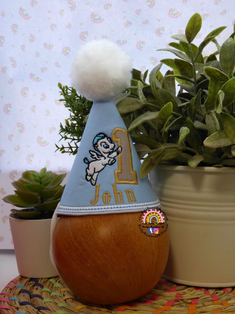 May include: A light blue birthday party hat with a white pom-pom on top. The hat features an embroidered design of a winged horse, the number 1, and the name "John" in gold thread. The hat is placed on a wooden form.