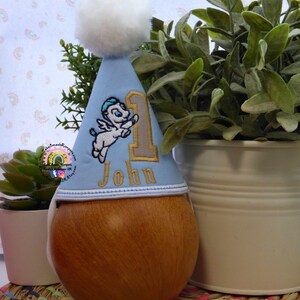 May include: A light blue birthday party hat with a white pom-pom and the number "1". The hat features an embroidered cartoon Pegasus and the name "John". The hat is placed on a wooden object.