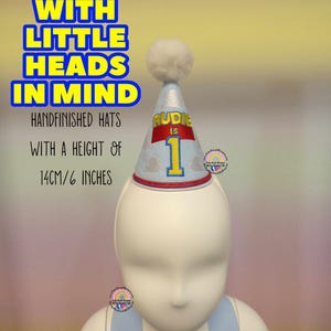 May include: A light blue and silver party hat with a white pom-pom on top. The hat is decorated with the text "RUDIE IS 1" and is 6 inches tall. The text "MADE WITH LITTLE HEADS IN MIND" is also visible.