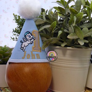 May include: A light blue birthday party hat with a white pom-pom, embroidered with a Pegasus and the number 1 in gold. The name "John" is embroidered below the number. The hat is displayed on a wooden form.