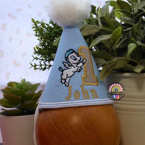 May include: A light blue birthday party hat with a white pom-pom on top. The hat features an embroidered cartoon Pegasus and the number 1 in gold. The name "John" is embroidered in gold below the number.