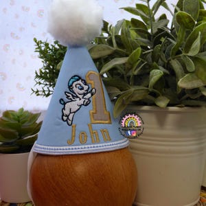 May include: A light blue birthday party hat with a white pom-pom and embroidered details. The hat features a cartoon Pegasus, the number 1, and the name "John" in gold thread. The hat is placed on a wooden base.