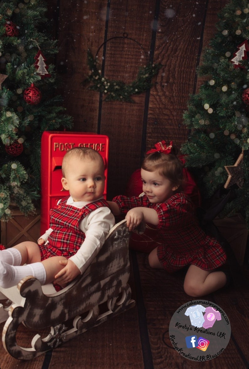 May include: Two toddlers wearing red and black plaid outfits pose for a Christmas photo. One child sits in a wooden sleigh while the other child reaches out to touch the sleigh. A red post office box is in the background.