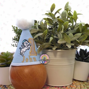 May include: A light blue birthday party hat with a white pom-pom and embroidered details. The hat features a cartoon angel, the number 1, and the name "John". The hat is placed on a wooden form, with plants in the background.