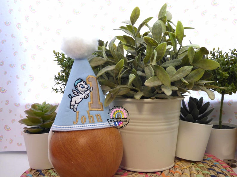 May include: A light blue birthday party hat with a white pom-pom and embroidered details. The hat features a cartoon angel, the number 1, and the name "John". The hat is placed on a wooden form, with plants in the background.
