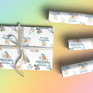 May include: White wrapping paper with a repeating pattern of cartoon characters, clouds, and the text "Happy Birthday Jermaine" and "ONEder Boy". A gift is wrapped with a white ribbon. Rolls of matching paper are also visible.