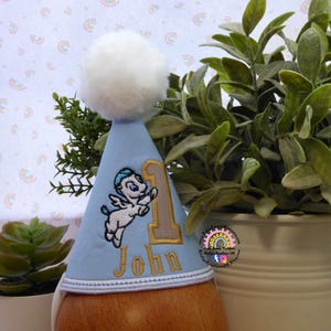 May include: A light blue birthday party hat with a white pom-pom on top. The hat features an embroidered design of a winged creature, the number 1, and the name "John" in gold thread. The hat is designed for a first birthday celebration.