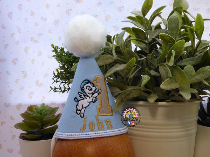 May include: A light blue birthday party hat with a white pom-pom on top. The hat features an embroidered design of a winged creature, the number 1, and the name "John" in gold thread. The hat is designed for a first birthday celebration.