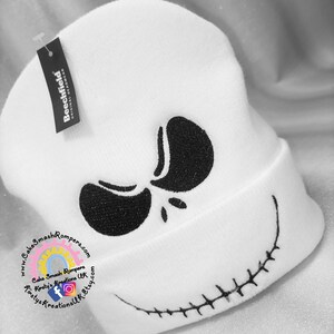 May include: White knit beanie with a black embroidered Jack Skellington face. The beanie has a stitched smile and the eyes are partially closed.