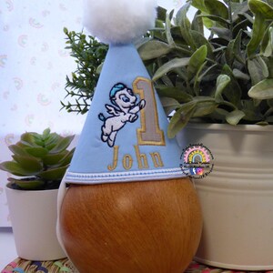 May include: A light blue birthday party hat with a white pom-pom, embroidered with a cartoon Pegasus and the number 1. The name "John" is embroidered in gold thread. The hat is displayed on a wooden form.