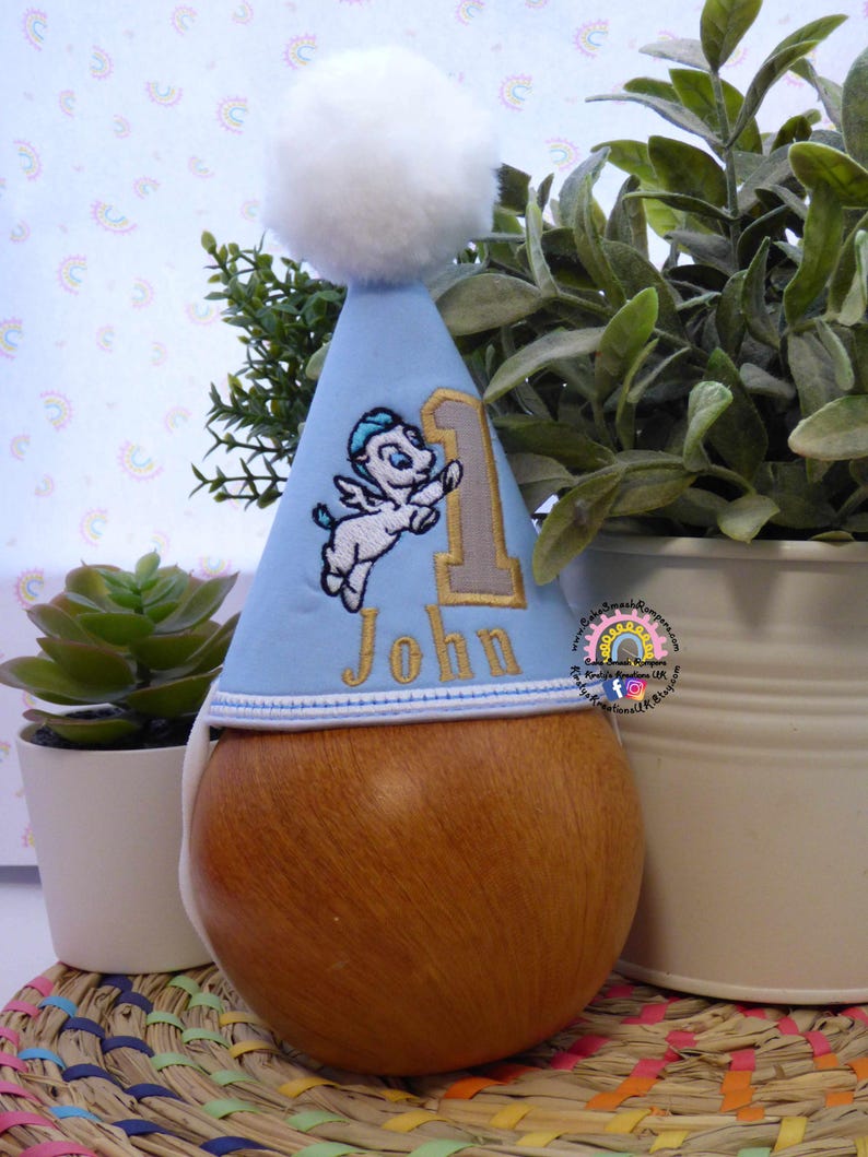 May include: A light blue birthday party hat with a white pom-pom, embroidered with a cartoon Pegasus and the number 1. The name "John" is embroidered in gold thread. The hat is displayed on a wooden form.