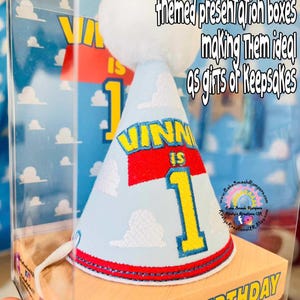 May include: A light blue birthday party hat with the name "VINNI IS 1" in yellow and red, and a white pom-pom. The hat is in a clear box with the words "HAPPY BIRTHDAY" on the box.