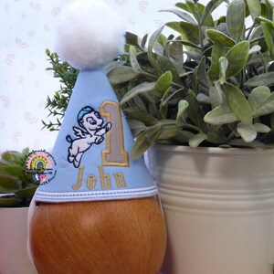 May include: A light blue birthday party hat with a white pom-pom, featuring an embroidered cartoon Pegasus and the name "John" with the number "1". The hat is placed on a wooden object, with greenery in the background.
