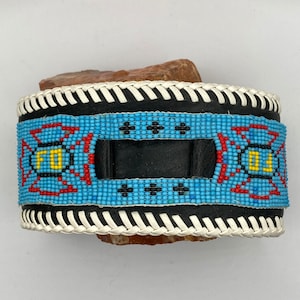 Puede incluir: Un brazalete de cuero con cuentas, con fondo azul, detalles rojos y amarillos y una correa de cuero negro. El brazalete tiene un ribete trenzado blanco.