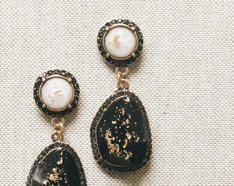 May include: A pair of black and gold earrings with a white pearl-like center. The earrings have a teardrop shape with a black stone border and gold flecks.