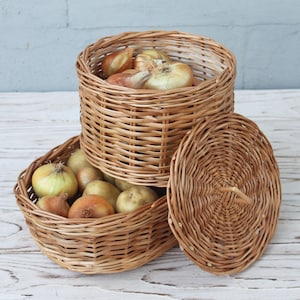 May include: Three woven wicker baskets, two round and one oval, filled with onions. The baskets are light brown and sit on a white wooden surface. One round basket has a lid. The onions are various sizes and colors.