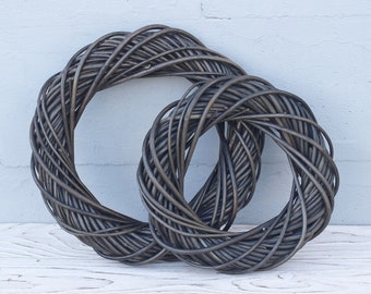 May include: Two black woven wreaths, one larger than the other, are displayed against a white brick wall. The wreaths are made of natural materials and have a rustic look.