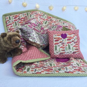 Op de afbeelding: Een bruine teddybeer zit op een roze en groene patchworkdeken met een bosprint. De deken heeft een groene rand en een roze stippenpatroon. Een klein kussen met een roze stippenpatroon en een paars hart ligt op de deken.